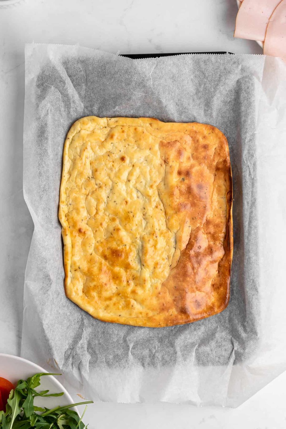 A freshly baked rectangular cottage cheese flatbread on parchment paper, resting on a marble surface. Ingredients like arugula, tomatoes, and slices of ham are partially visible around the quiche.