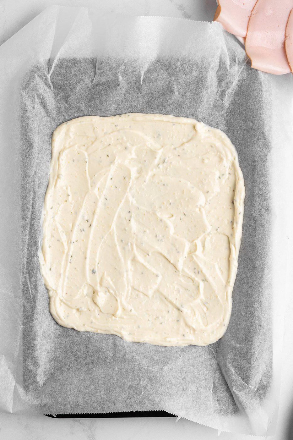 A baking tray lined with parchment paper holds an even spread of creamy, seasoned dough or batter. The surface is smooth and speckled with herbs. A few slices of deli meat are visible in the corner on a light background.
