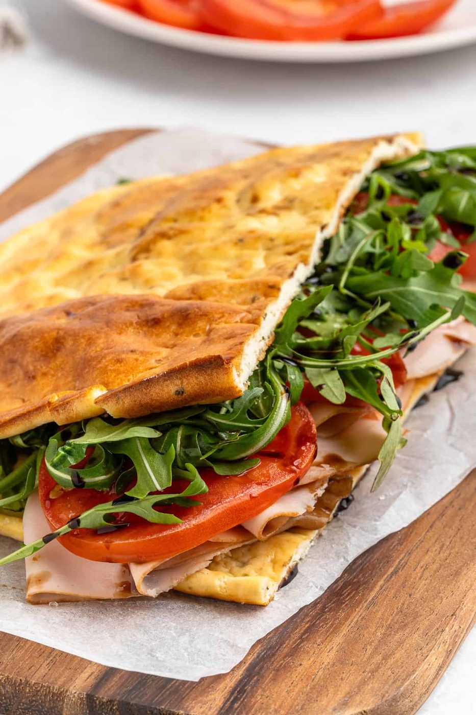 A sandwich on a wooden board, made with flatbread, filled with arugula, sliced tomatoes, and deli turkey. The flatbread is folded over the ingredients, which are visible. A plate with more tomato slices is in the background.
