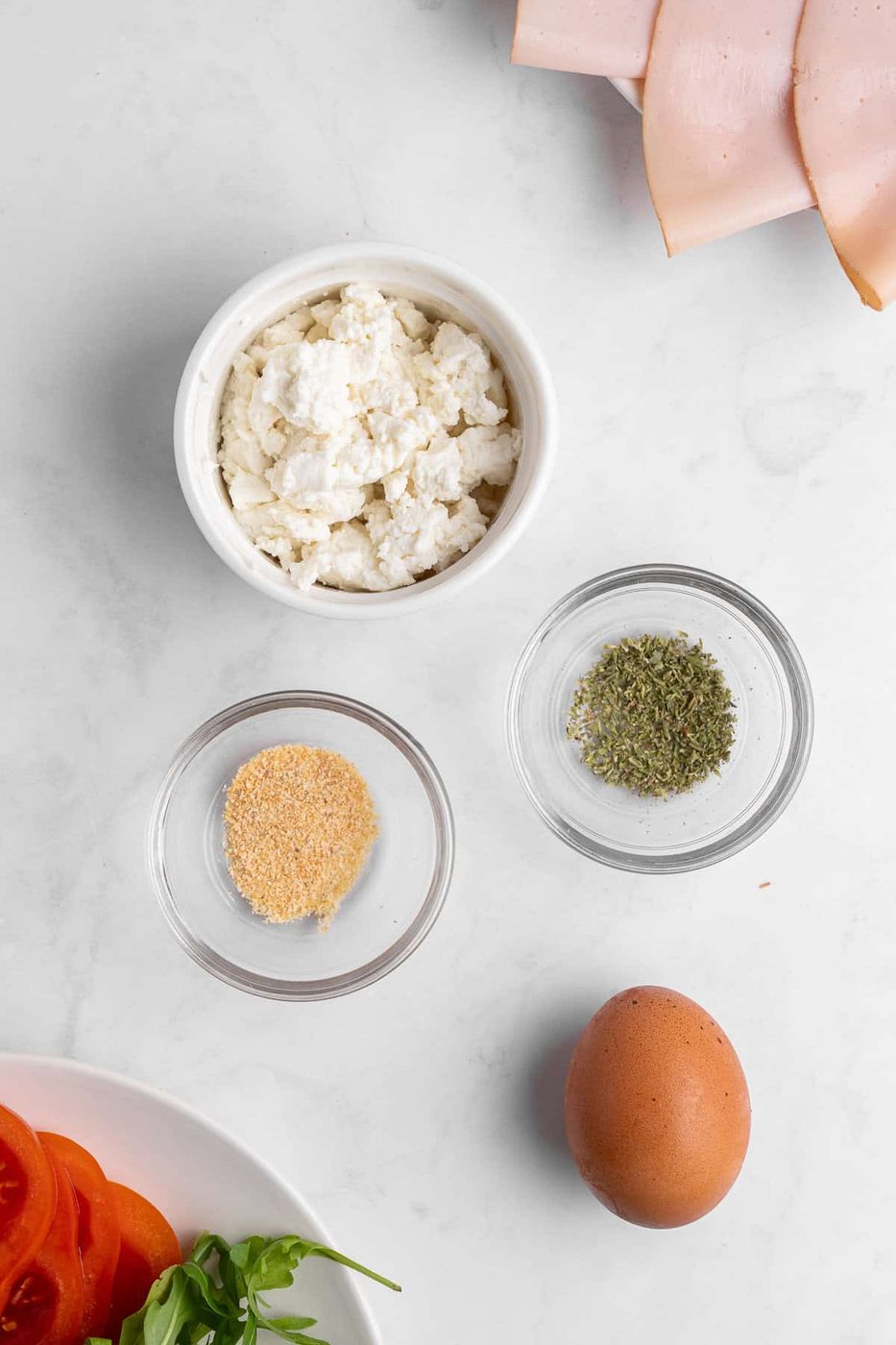 Top-down view of ingredients on a white surface: a bowl of ricotta cheese, dried herbs, garlic powder, sliced ham, a brown egg, fresh tomato slices, and arugula leaves.