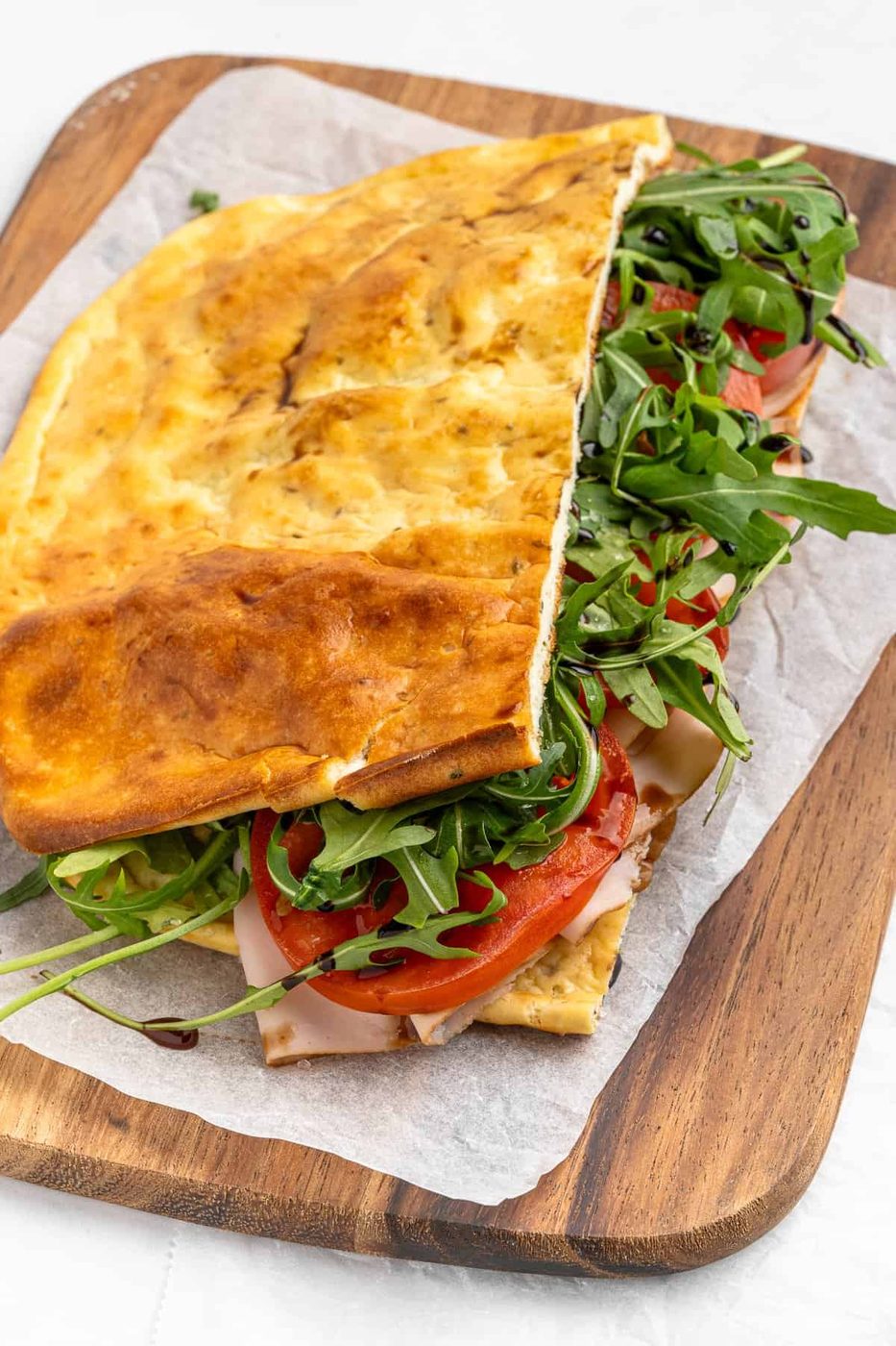 A folded flatbread sandwich on a wooden board, filled with sliced turkey, fresh tomato, arugula, and cheese. The bread appears golden and lightly toasted, resting on a piece of parchment paper.