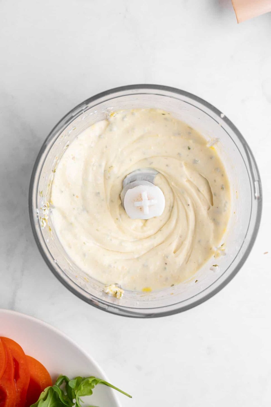 A food processor containing creamy, light-colored dressing with visible herbs. Next to it is a plate with sliced tomatoes, and a few leaves of fresh basil on a white marble surface.