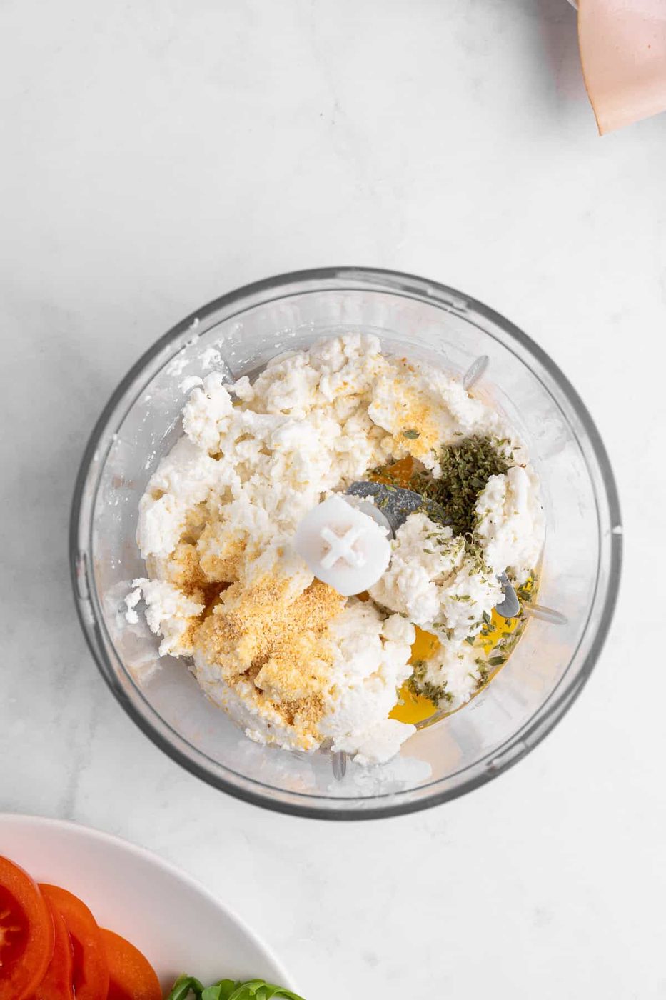 A food processor filled with ricotta cheese, breadcrumbs, herbs, and seasoning. Below, a plate with sliced tomatoes and basil is partially visible. The background is a white marble surface.