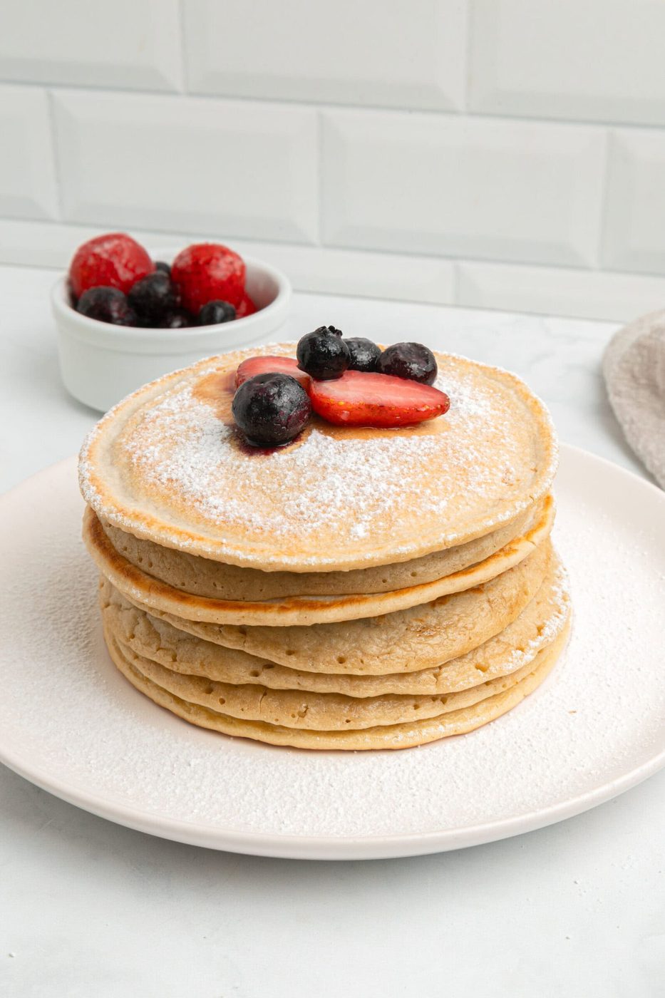 A stack of pancakes topped with powdered sugar, strawberries, and blueberries on a white plate, with a small bowl of mixed berries in the background.