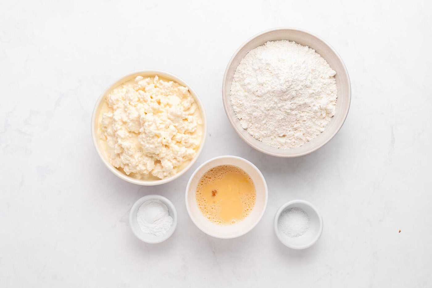 Five bowls on a white surface: one with cottage cheese, one with flour, one with beaten egg, one with baking powder, and one with salt.