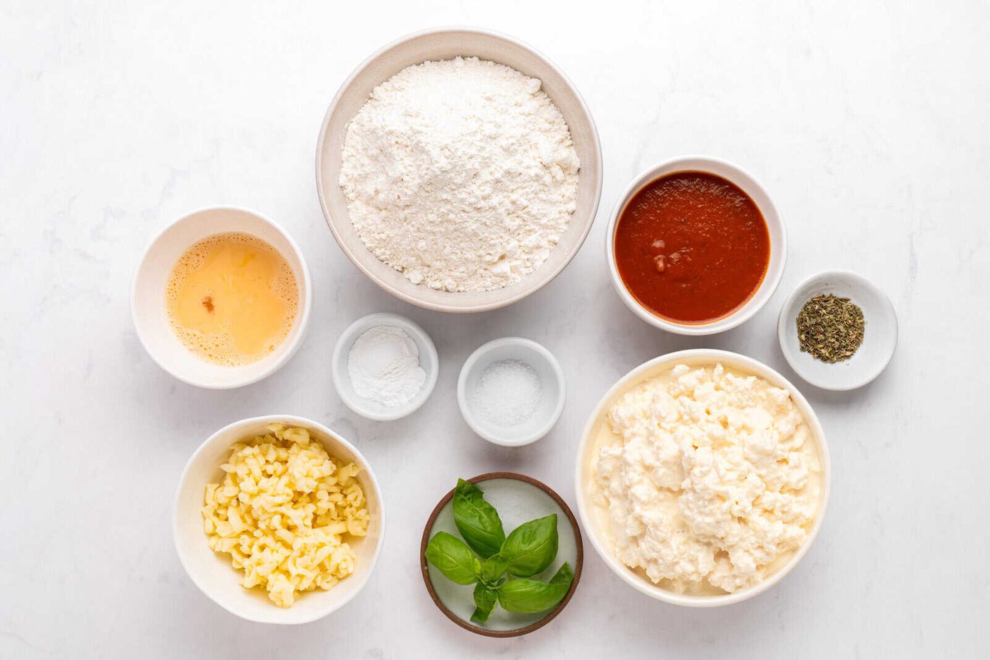 Various pizza ingredients in bowls on a white surface, including flour, tomato sauce, shredded cheese, ricotta cheese, beaten egg, baking powder, salt, dried herbs, and fresh basil leaves.