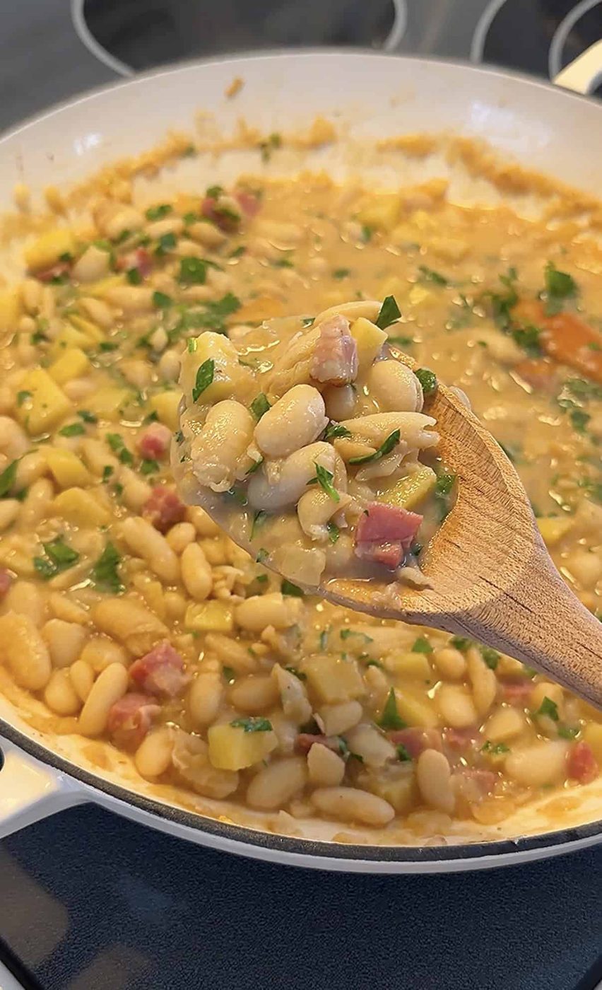 A wooden spoon holds up a serving of Creamy Bean & Potato Stew with chopped herbs and diced ham over a pot filled with the same creamy mixture.