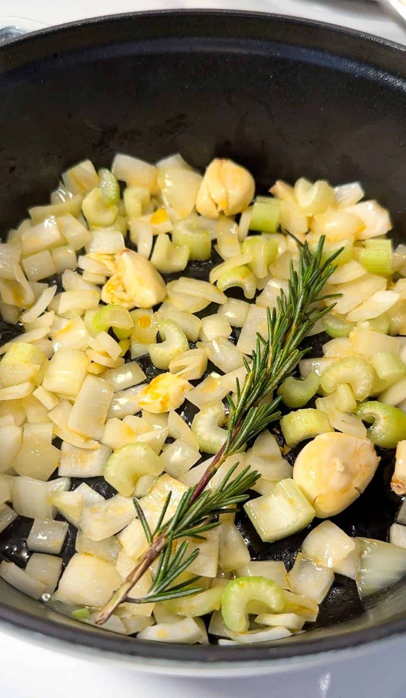 Chopped onions, celery, and garlic cloves are being sautéed in a black pan with a sprig of fresh rosemary.