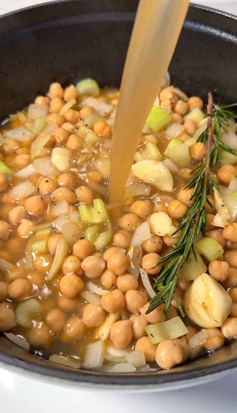 A black pot filled with chickpeas, chopped onions, and celery is being poured with a light broth. A sprig of rosemary and cloves of garlic are visible among the ingredients.
