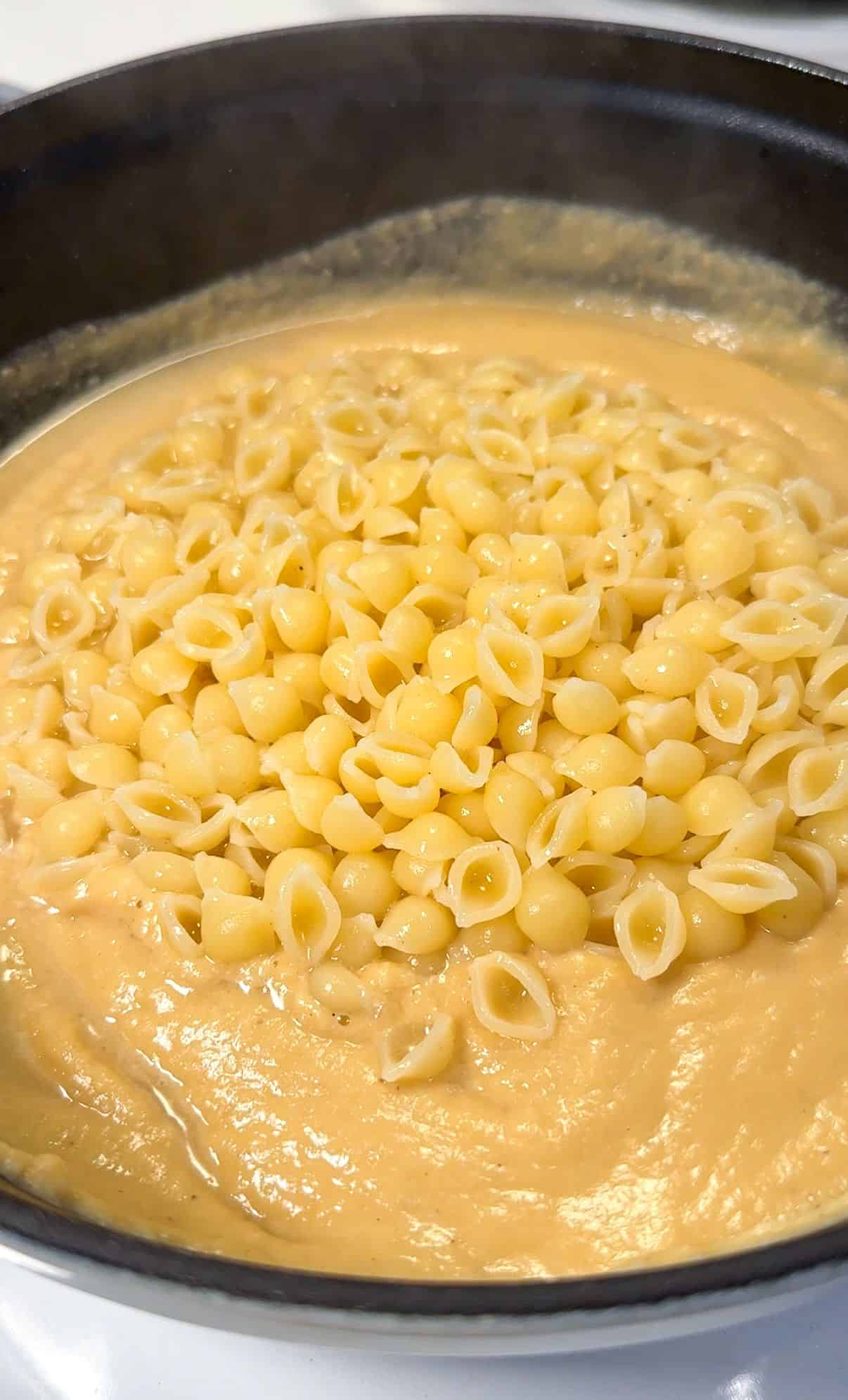 A black pan filled with creamy cheese sauce and small shell pasta. The pasta is partially mixed into the thick, smooth sauce, creating a rich, cheesy dish.