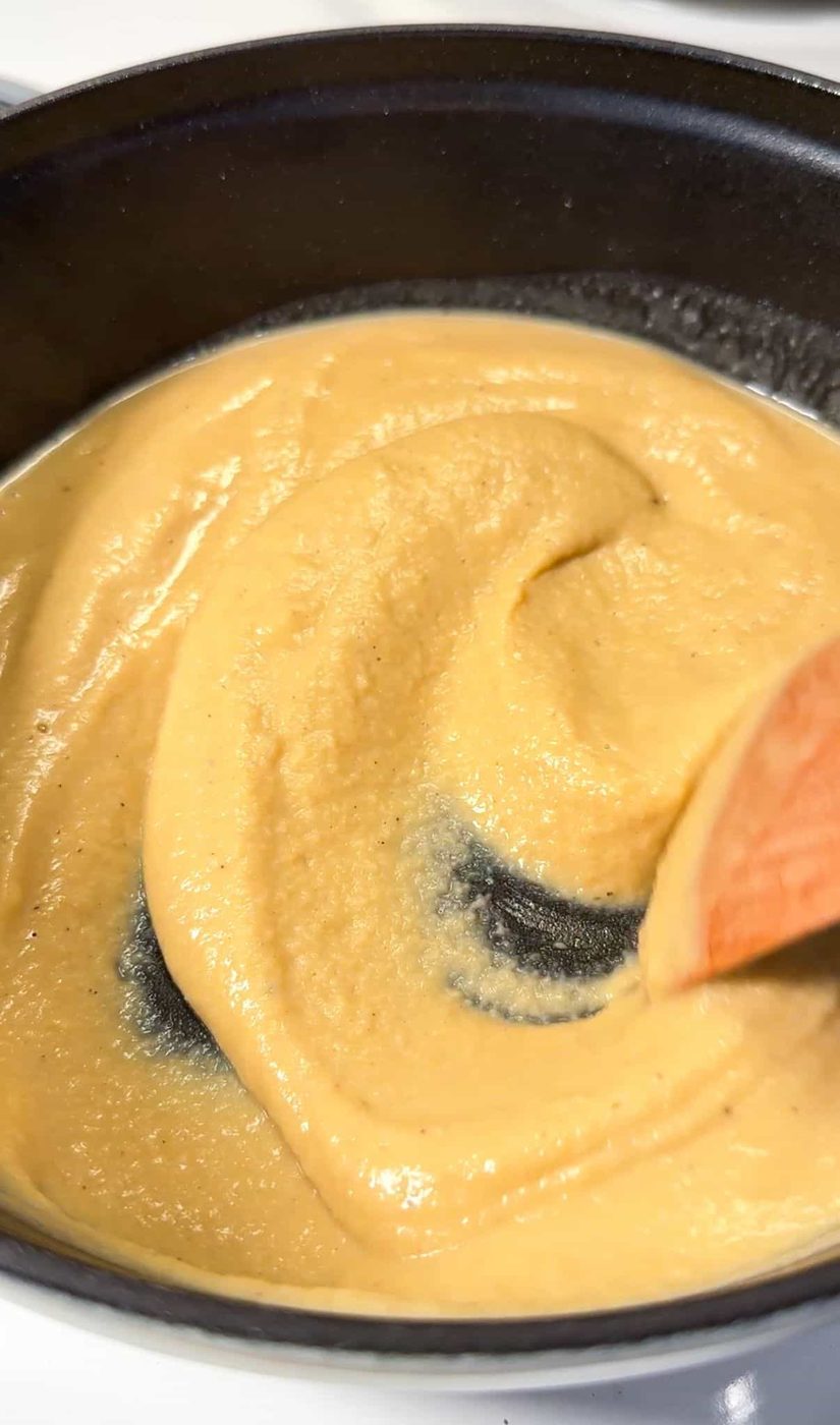 A close-up of a creamy, light brown sauce or batter being stirred in a black frying pan with a wooden spatula. The mixture appears smooth and thick, with a slight sheen on its surface.
