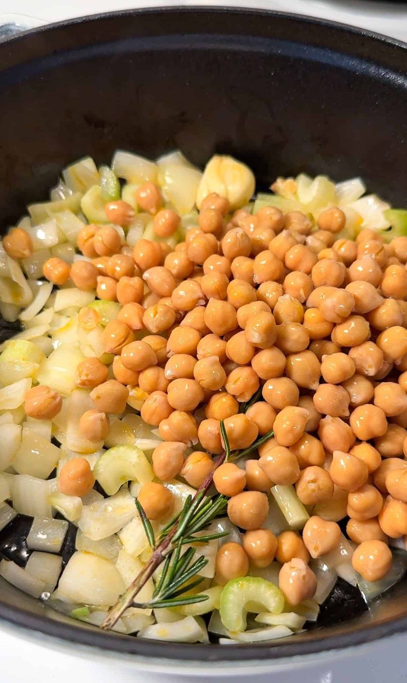 A black pot containing chopped onions, celery, garlic, and chickpeas. There's a sprig of rosemary on top, and the mixture appears to be ready for cooking.