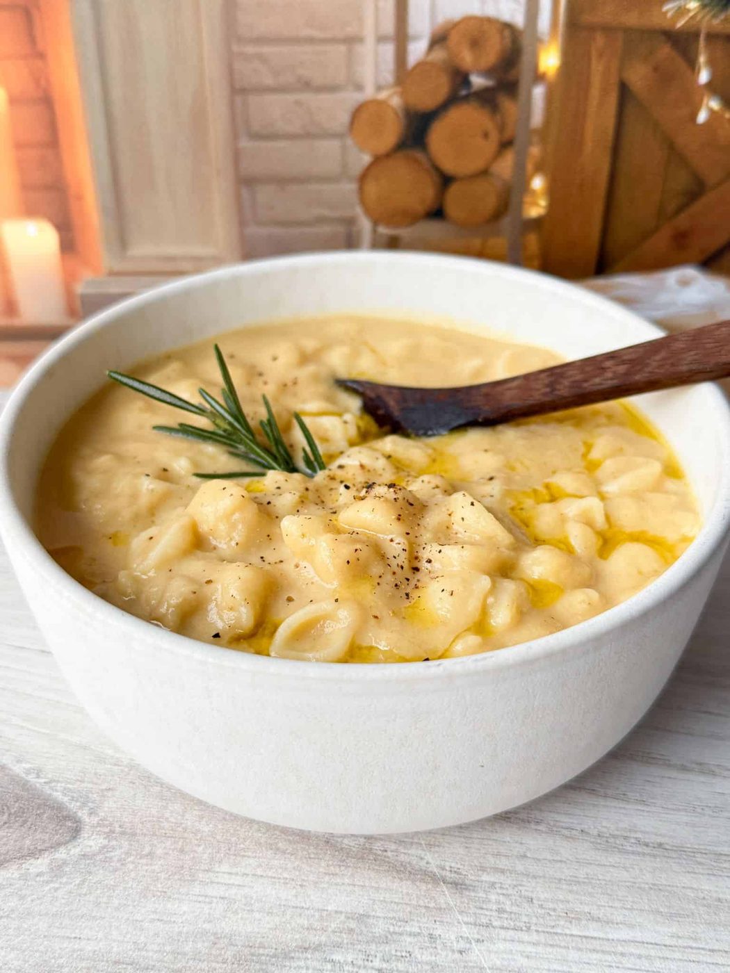 A bowl of creamy pasta soup garnished with a sprig of rosemary and freshly cracked black pepper, with a wooden spoon resting inside. The setting is cozy, featuring stacked wooden logs and a lit candle in the background.