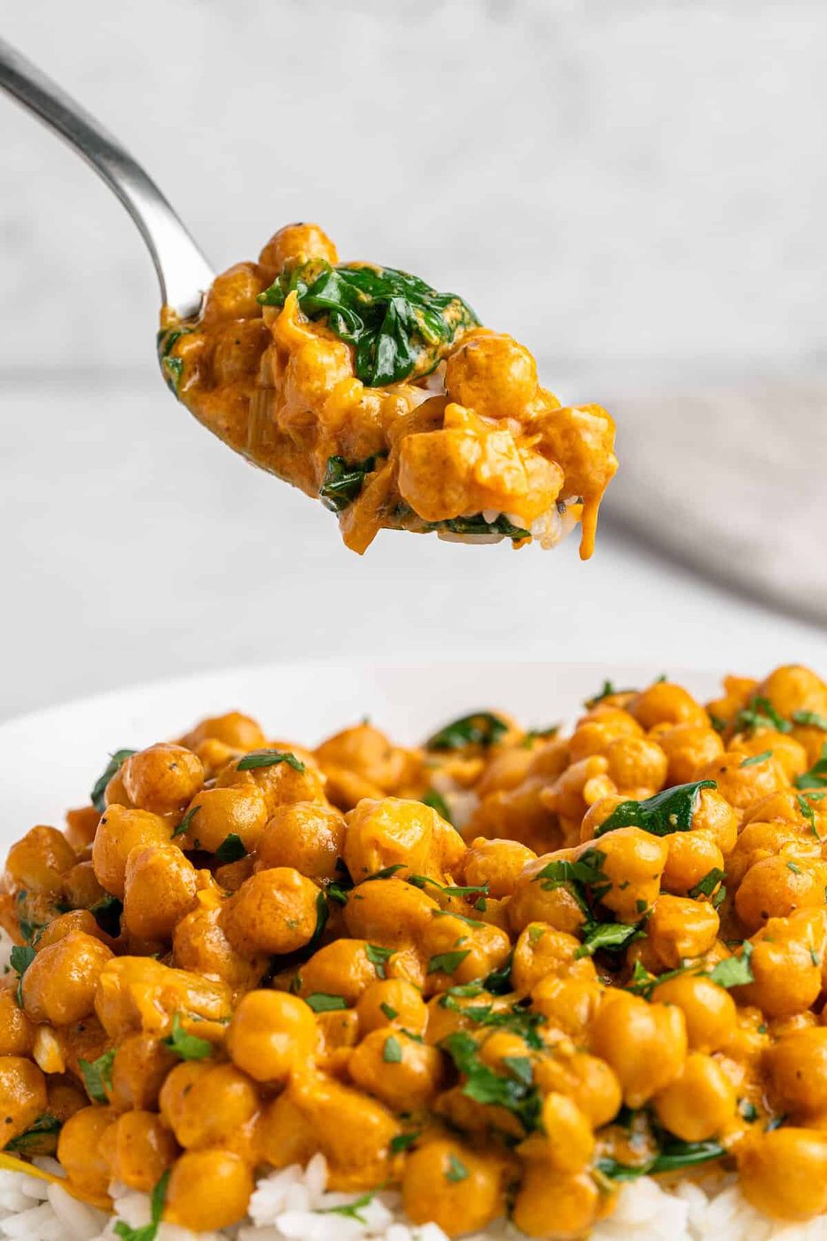 A spoonful of creamy chickpea curry with spinach is held above a plate. The curry is served on a bed of rice, with a rich, golden sauce and garnished with fresh herbs.