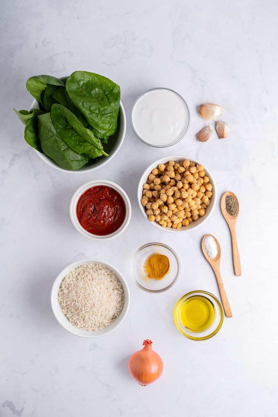 Ingredients laid out on a white surface like: fresh spinach, garlic cloves, tomato paste, uncooked rice, onion, and small bowls of olive oil, turmeric, pepper, and salt.