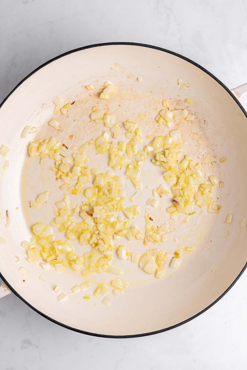 Chopped onions sautéing in a white pan on a marble surface. The onions are slightly golden, indicating they are being cooked. The pan shows hints of browning, suggesting the onions are being caramelized.
