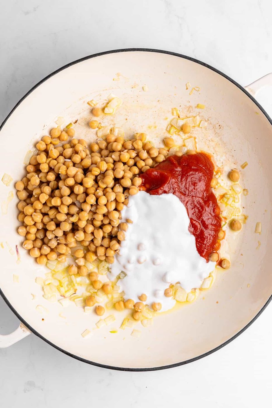 A white pot containing cooked chickpeas, chopped onions, tomato sauce, and coconut milk, ready to be mixed.