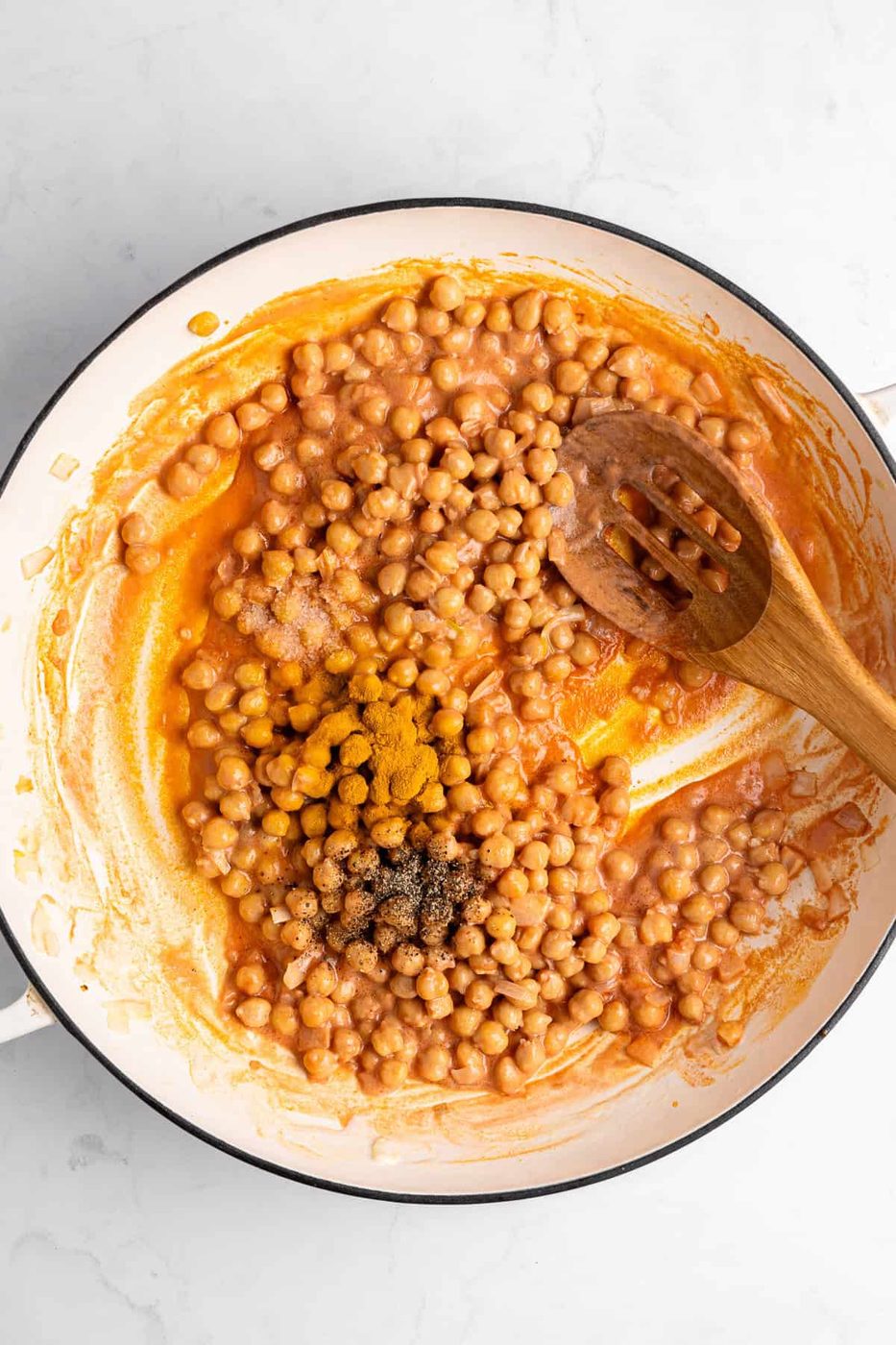 A skillet filled with garbanzo beans in a rich sauce, stirred with a wooden spoon. Seasonings including black pepper are visible on top. The background is a white marbled surface.