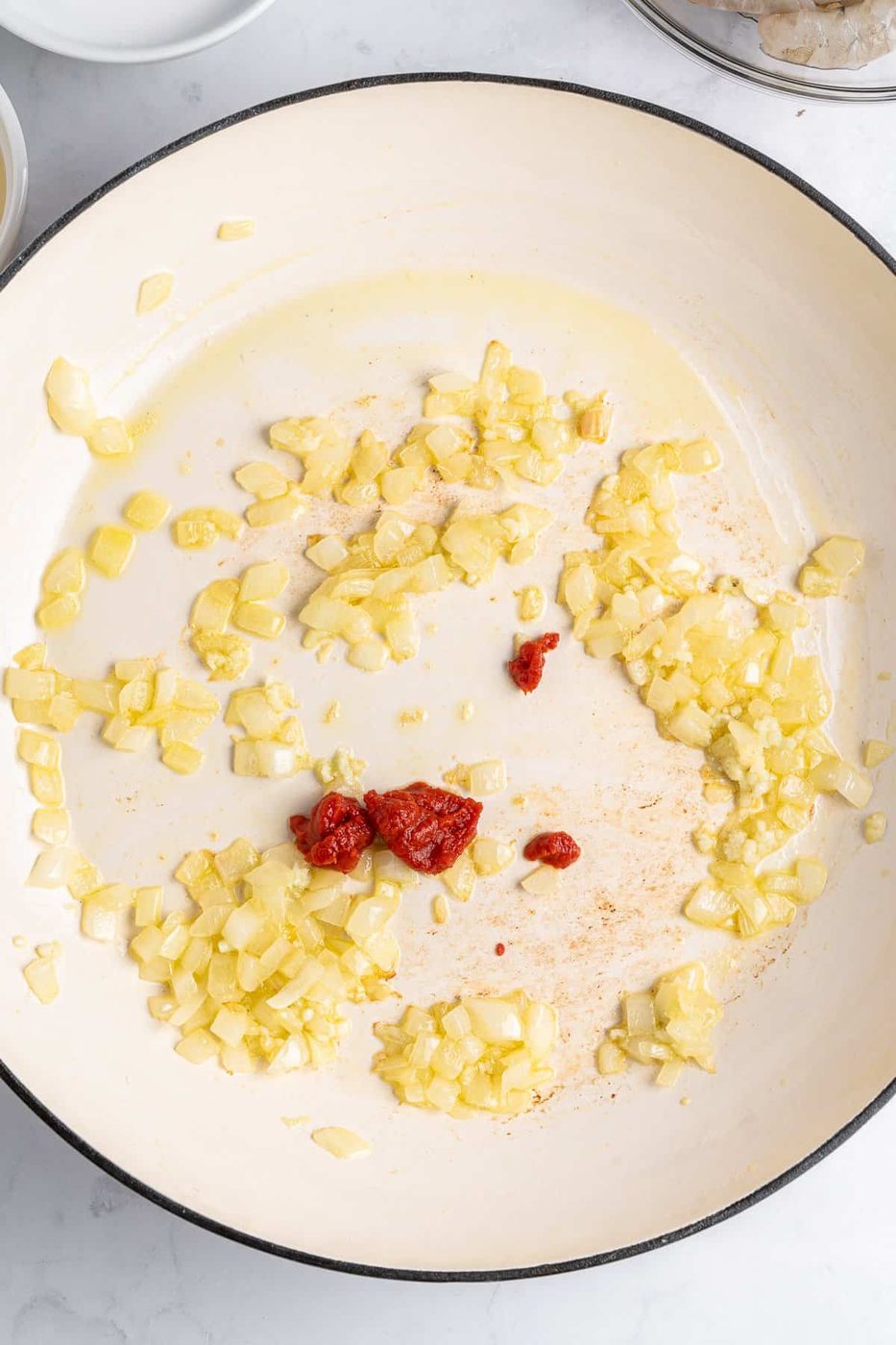 Chopped onions and garlic sautéing in a white pan with a small amount of tomato paste in the center. The ingredients are lightly cooked and glistening in oil. The pan sits on a white marble surface.