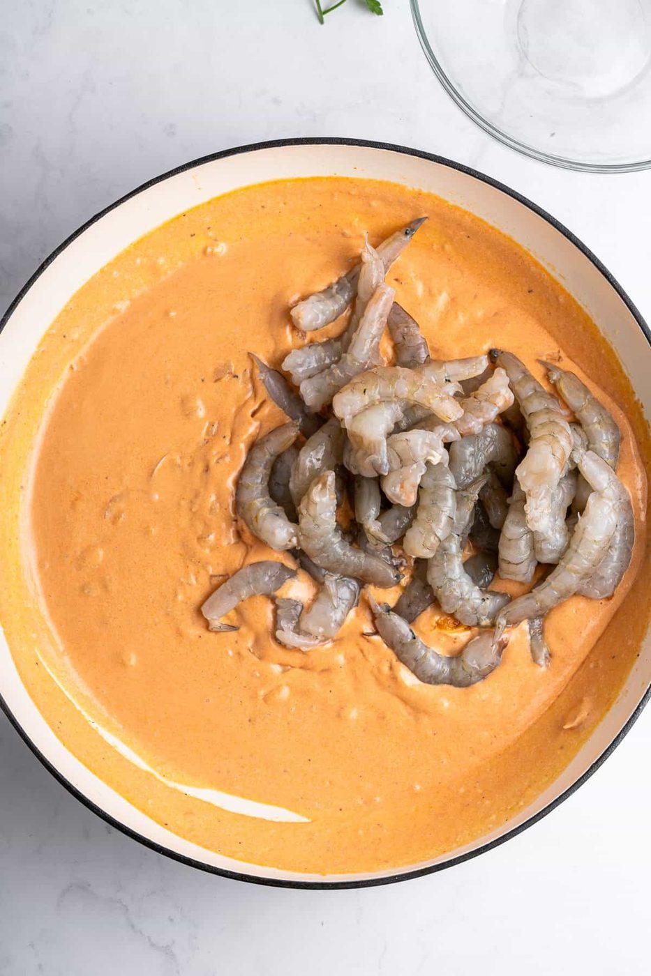 A skillet with a creamy sauce and uncooked shrimp on top. The sauce is smooth and rich, contrasting with the raw, gray shrimp. The skillet rests on a white marble surface, partially showing a glass bowl in the corner.