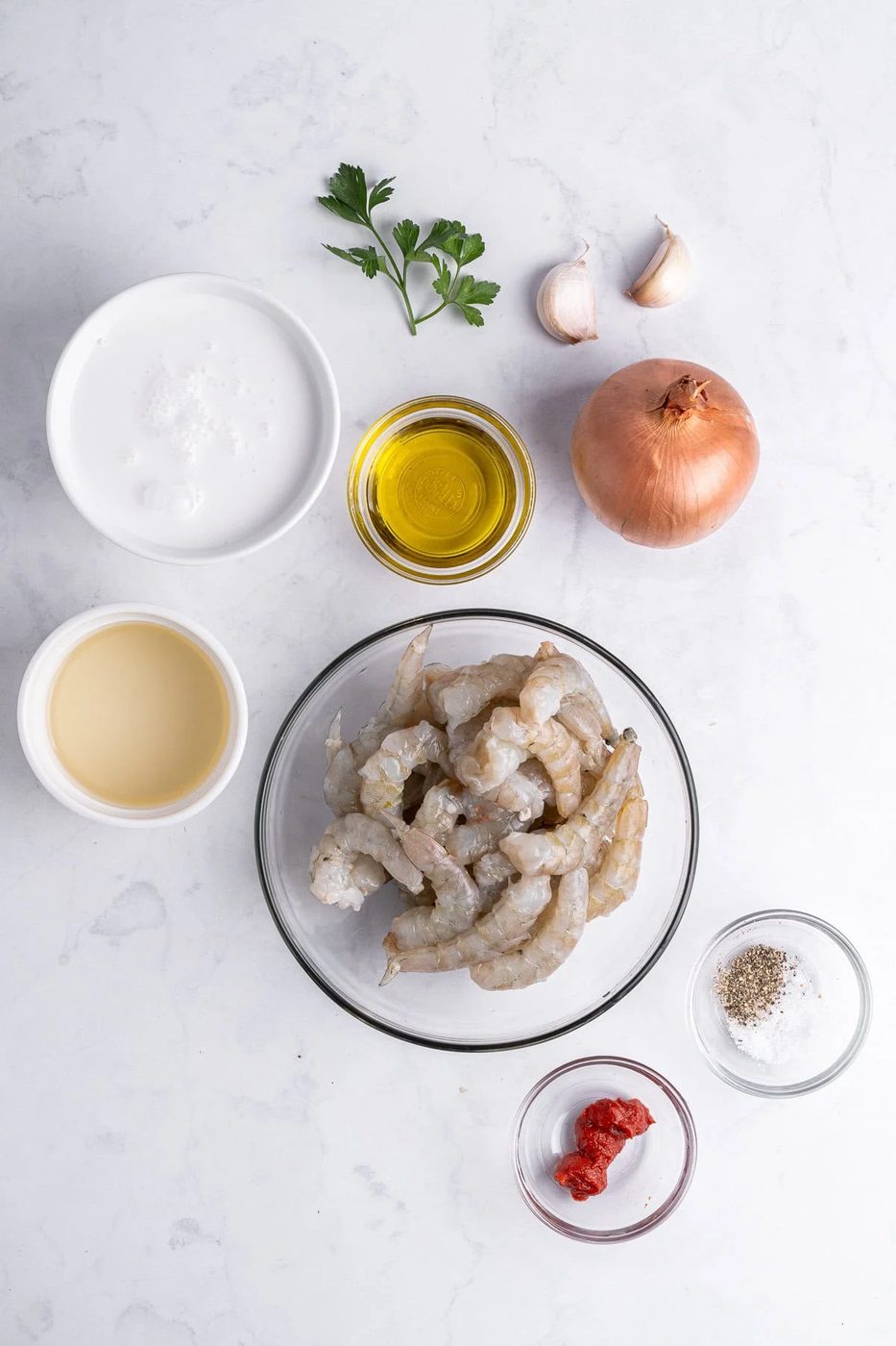 Top-down view of cooking ingredients on a white surface, including raw seafood in a bowl, an onion, garlic cloves, olive oil, coconut milk, white wine, tomato paste, parsley, salt, and pepper.