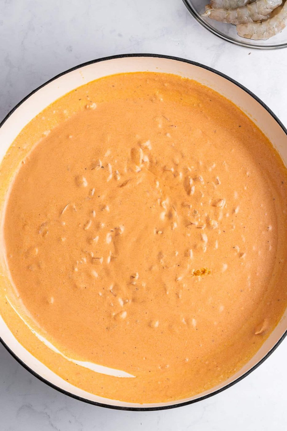 A pan filled with creamy sauce is placed on a white marble surface. The sauce has a slightly textured surface. In the upper corner, a bowl containing raw shrimp is partially visible.