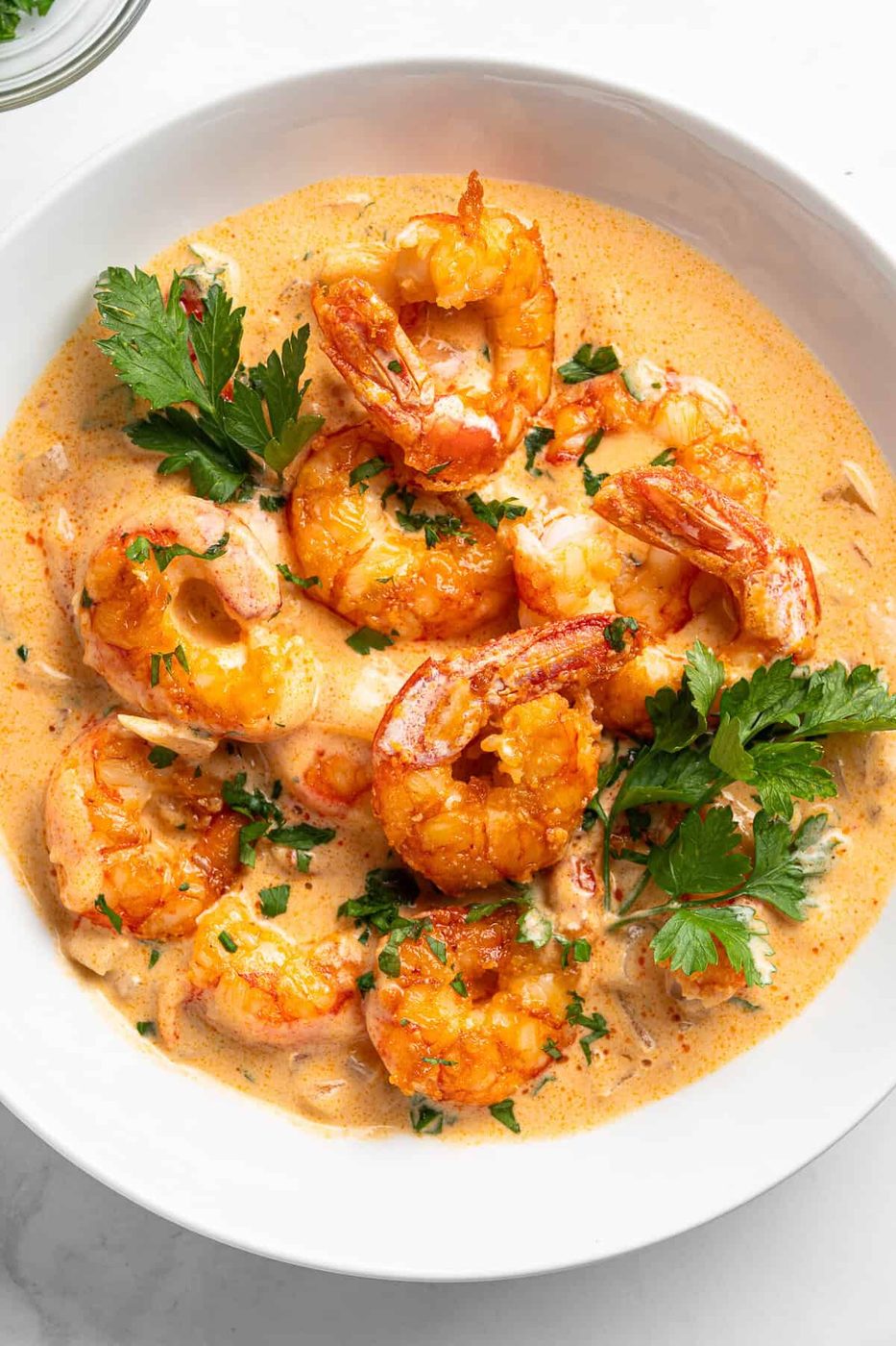 A white bowl filled with creamy garlic shrimp garnished with fresh parsley. The shrimp are cooked and have a golden color, nestled in a rich, orange-hued sauce. The dish is presented on a light background.