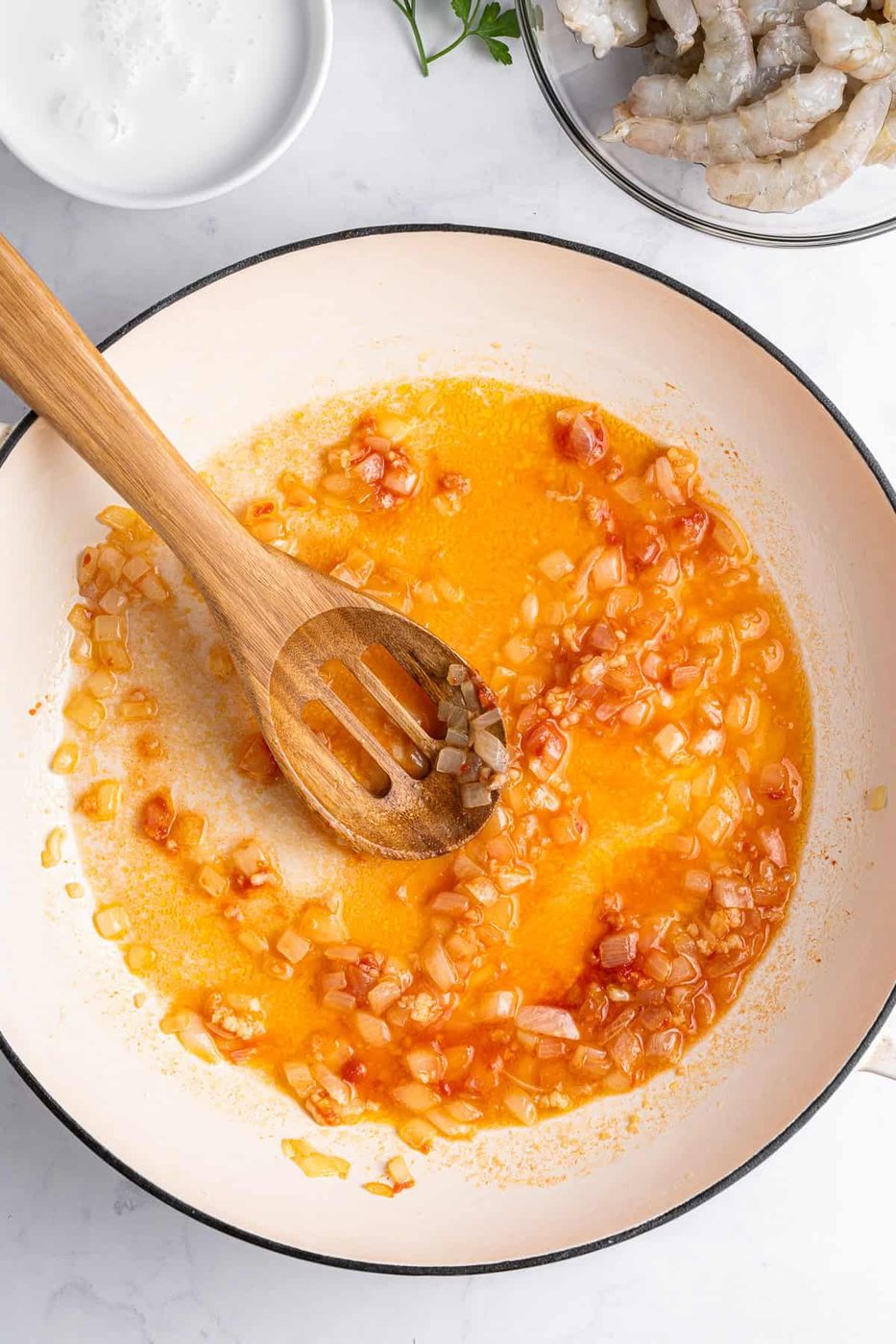 A pan with cooked onions and diced tomatoes in a reddish oil mixture, stirred by a wooden spoon. Nearby is a small bowl of white sauce. The scene is set on a white marble surface.