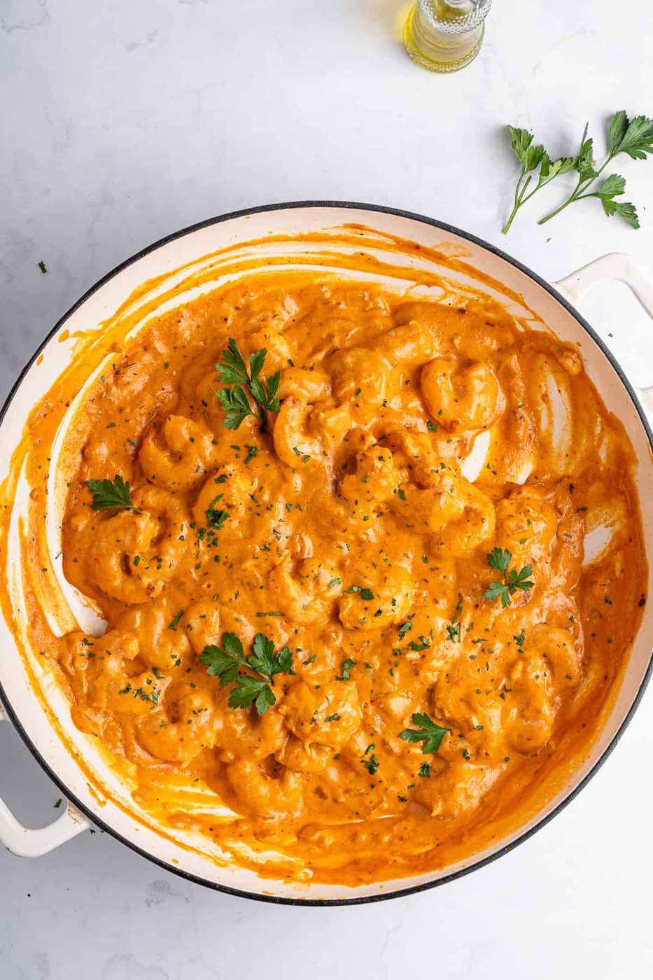 A white pan filled with creamy shrimp in a rich sauce, garnished with fresh parsley. The dish is set on a light marble surface with a sprig of parsley and a bottle of olive oil in the background.