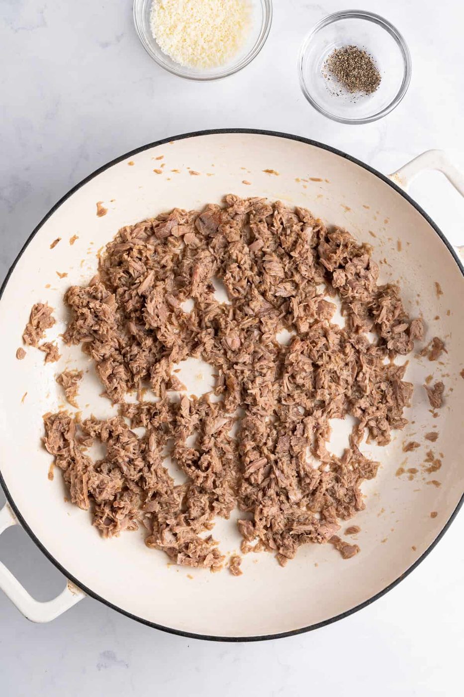 A white skillet on a marble surface contains cooked ground meat evenly spread across the bottom. Nearby, there are two small bowls, one with grated cheese and the other with ground black pepper.