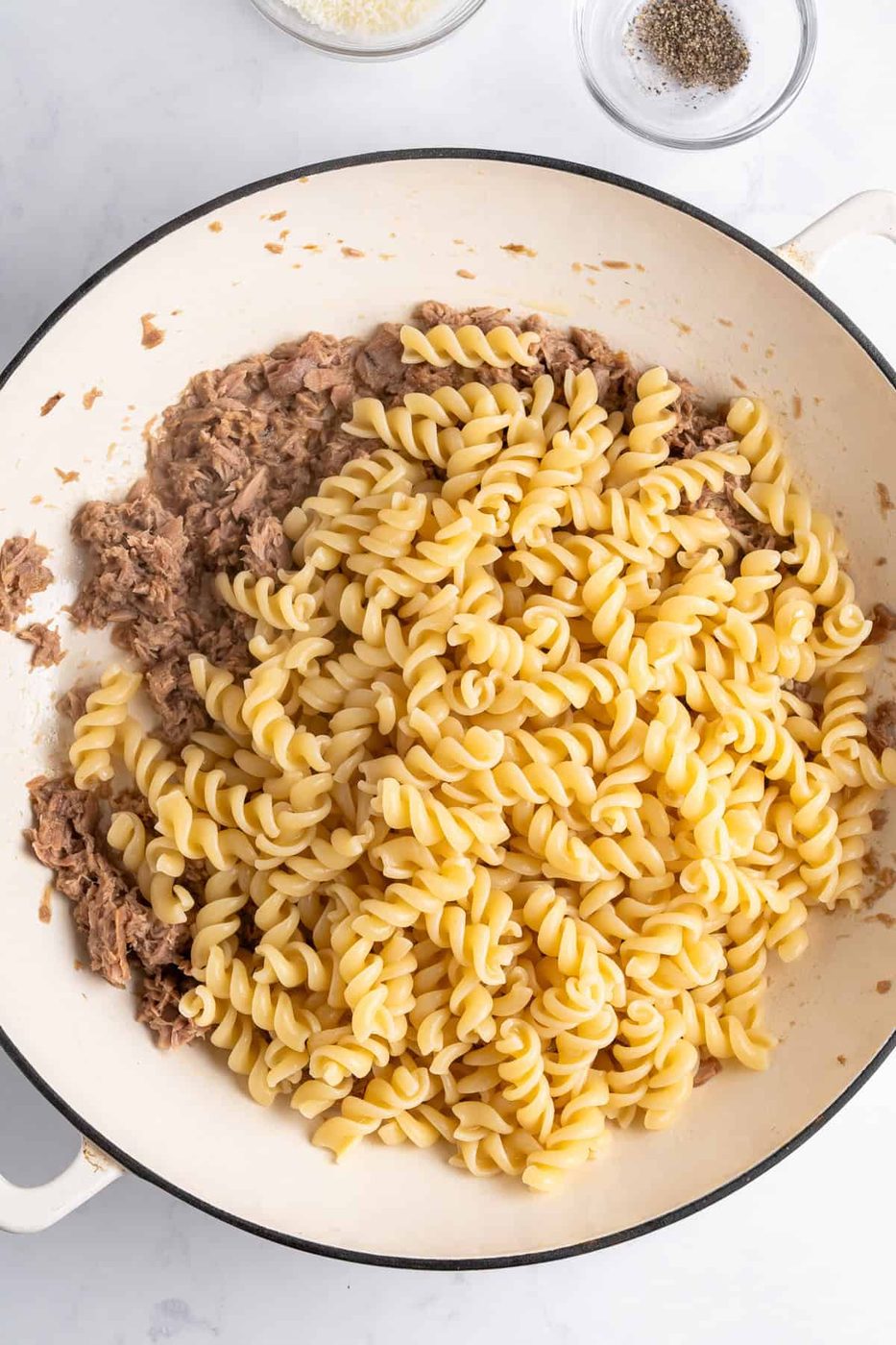 A white pan filled with fusilli pasta and tuna, ready to be mixed. Small bowls of grated cheese and black pepper are visible in the background on a white surface.