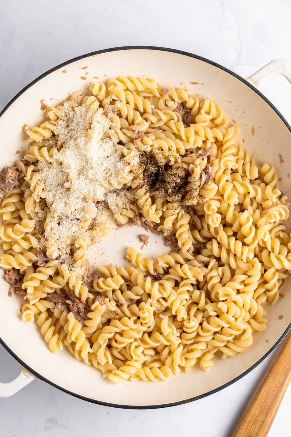 A white skillet filled with cooked fusilli, topped with grated cheese, black pepper, and small bits of meat. The ingredients are ready to be mixed for a final dish. The skillet has a white handle, and a wooden spoon is visible beside it.