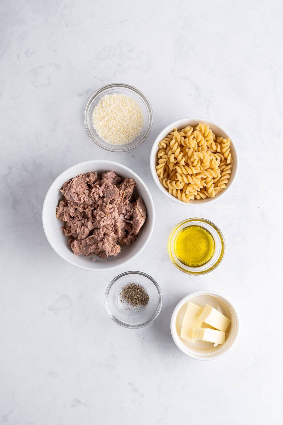 Ingredients on a white surface: a bowl of tuna, fusilli pasta, grated cheese, olive oil, butter cubes, and a bowl with salt and pepper.