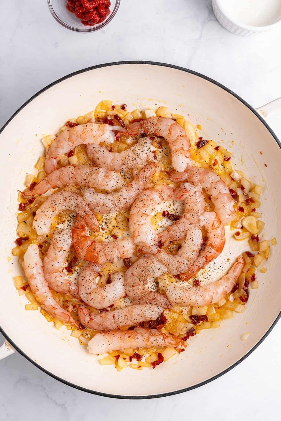 A pan filled with raw shrimp seasoned with pepper, resting on sautéed onions and diced sun-dried tomatoes. A small bowl of tomato paste is visible in the top left corner. The background is a white marble surface.