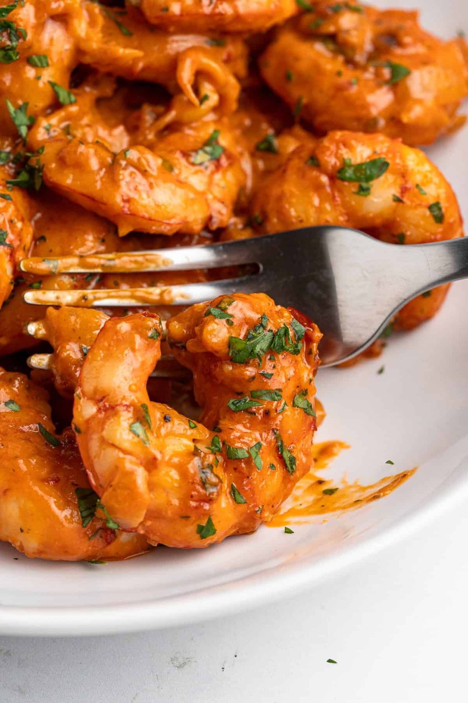 Close-up of a fork holding a creamy tuscan shrimp coated in a rich, creamy sauce with finely chopped herbs on a white plate. Other shrimp are visible in the background, all garnished with herbs.