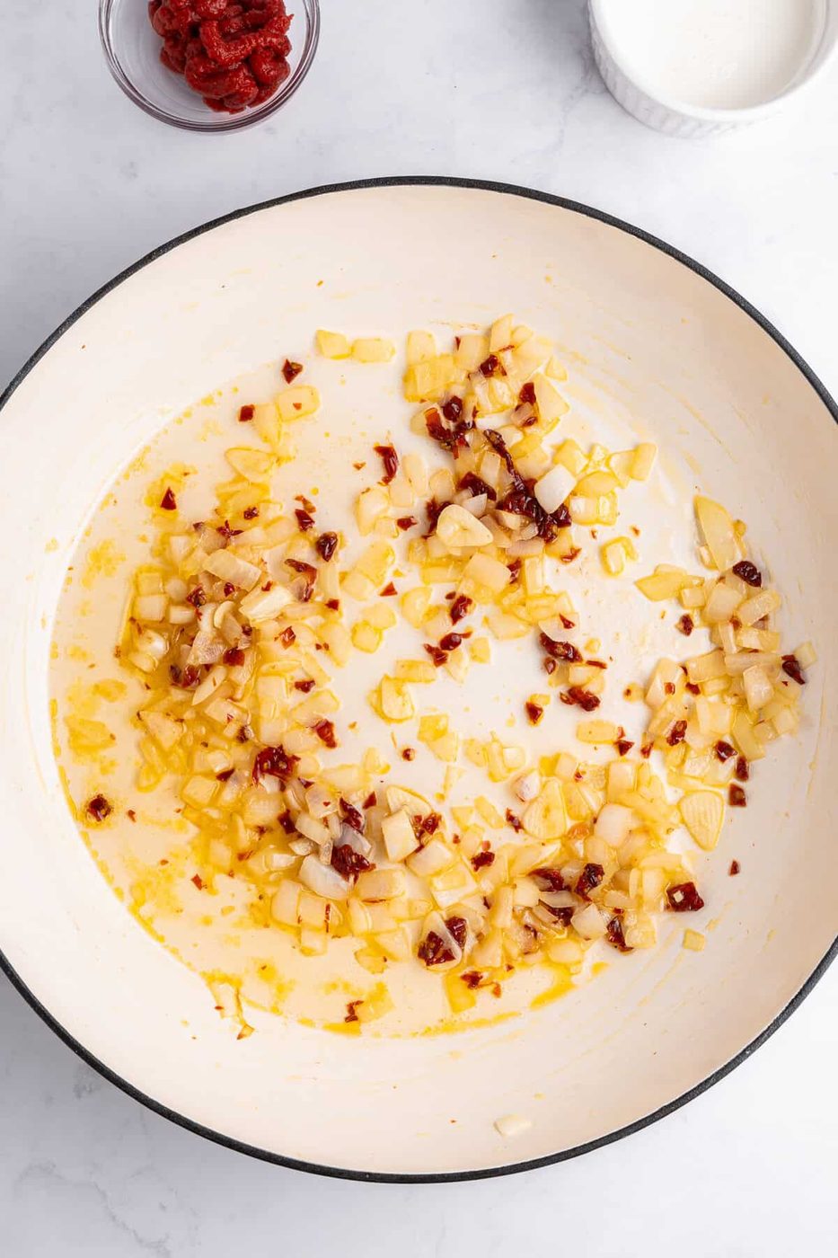Chopped onions and red pepper flakes sautéing in oil in a large white sauté pan. Bowls with ingredients, including tomato paste and cream, are placed nearby on a white marble surface.