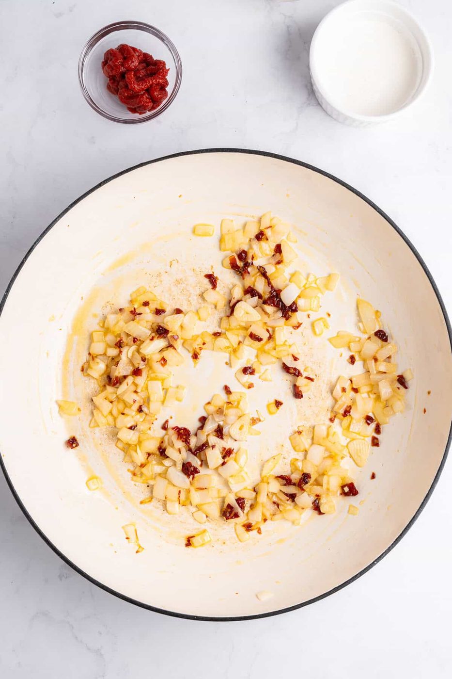 A white skillet on a marble surface contains sautéed onions and spices. Nearby are small bowls with tomato paste and cream.