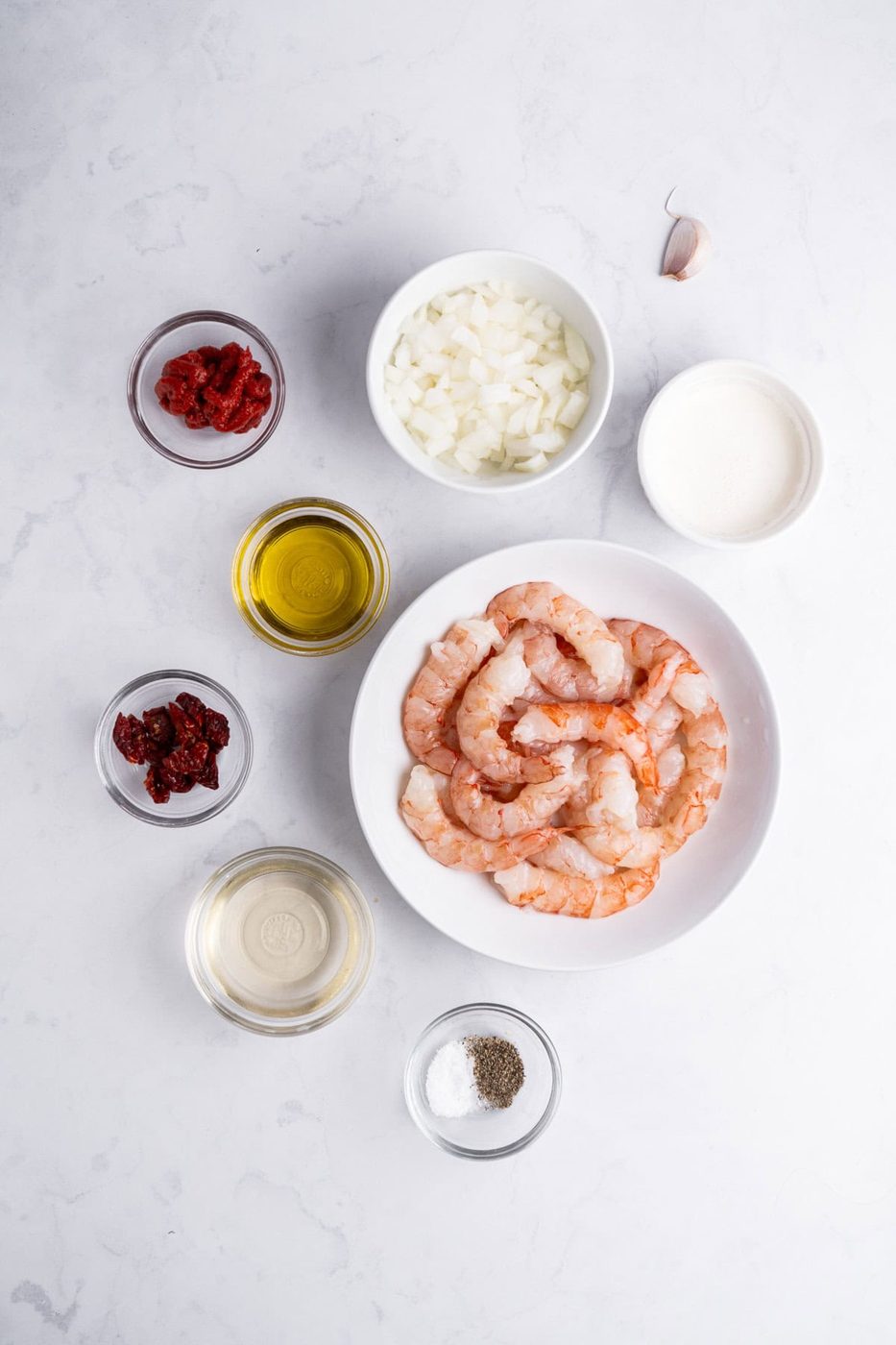 A flat lay of shrimp in a white bowl surrounded by bowls containing chopped onions, tomato paste, sun-dried tomatoes, olive oil, cream, white wine, a garlic clove, and a small dish of salt and pepper on a light marble surface.