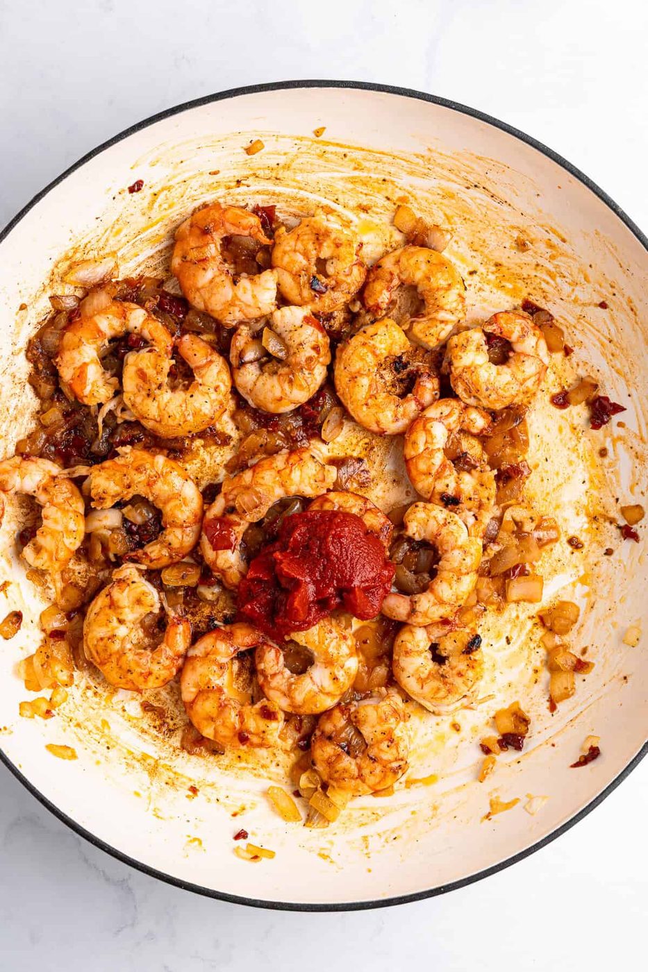 A white pan filled with cooked shrimp, chopped onions, and a blend of spices. A dollop of tomato paste is in the center, and everything is mixed together with visible seasonings. The background is a light, smooth surface.