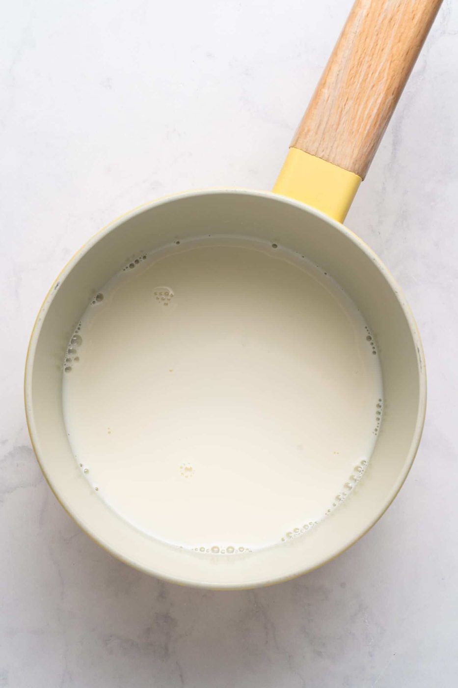 A small saucepan with a wooden handle, filled with milk, sits on a light marble surface.