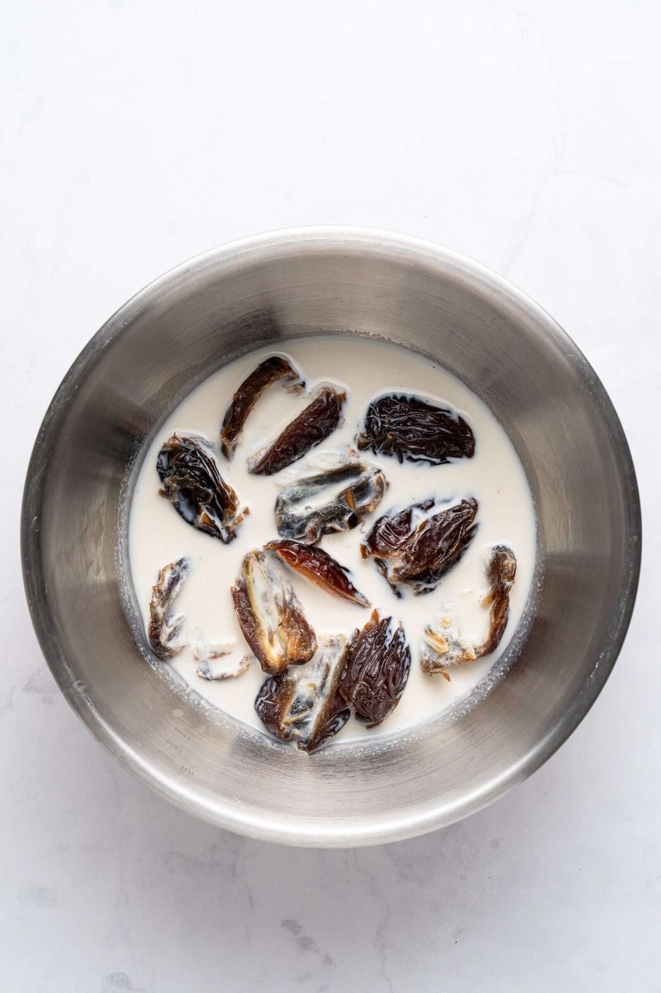 A stainless steel bowl contains morel mushrooms soaking in a light-colored liquid. The bowl is placed on a white marble surface.