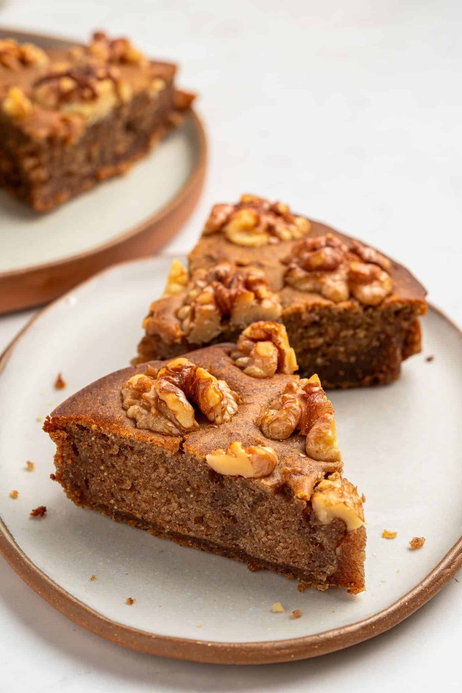 Two slices of walnut cake on a round plate, topped with whole walnuts. The cake has a rich brown color, and another plate with more cake slices is visible in the background. The setting is on a light-colored surface.