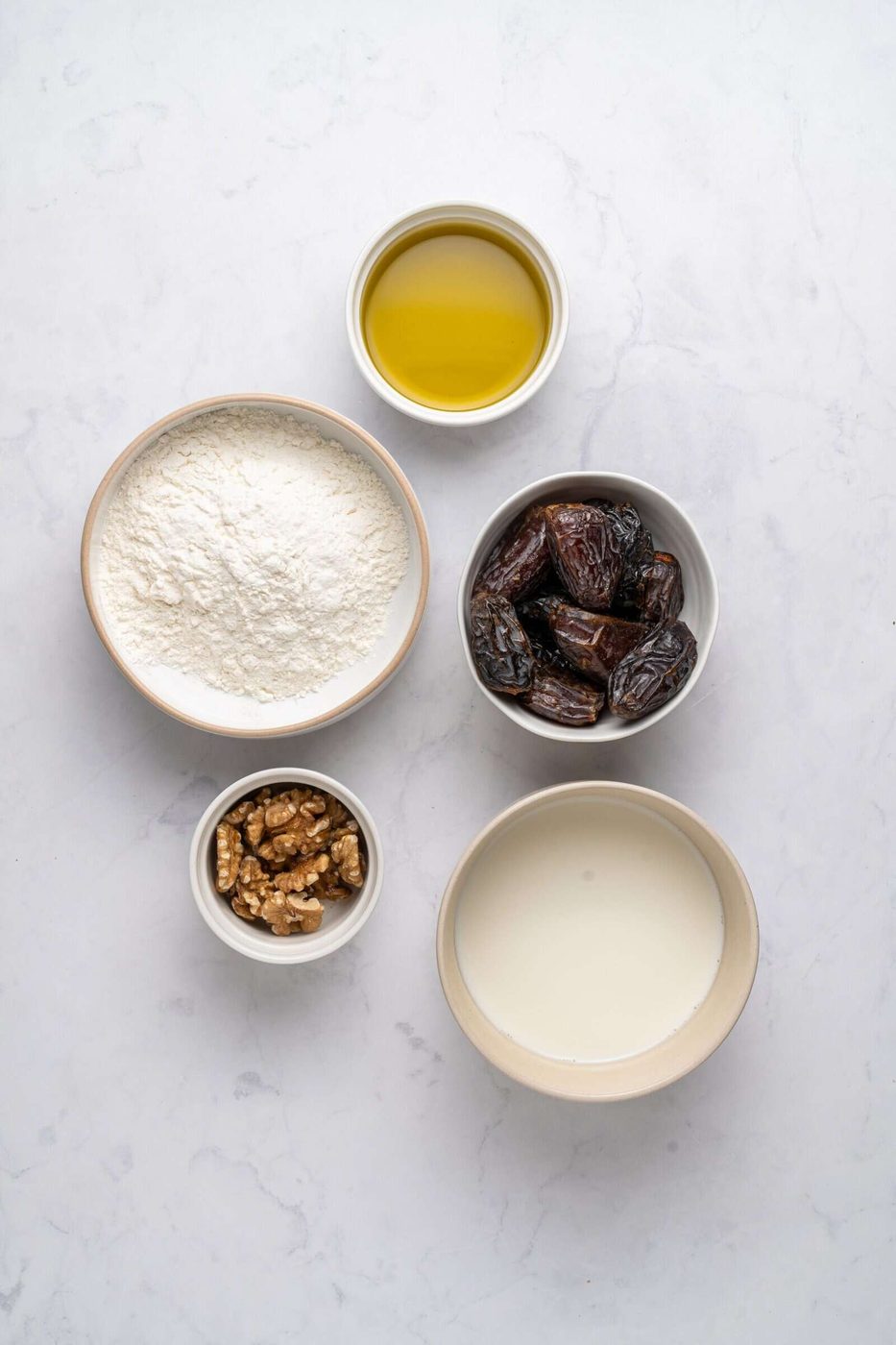 Bowls of ingredients on a marble surface: flour, olive oil, whole dates, milk, and walnuts.