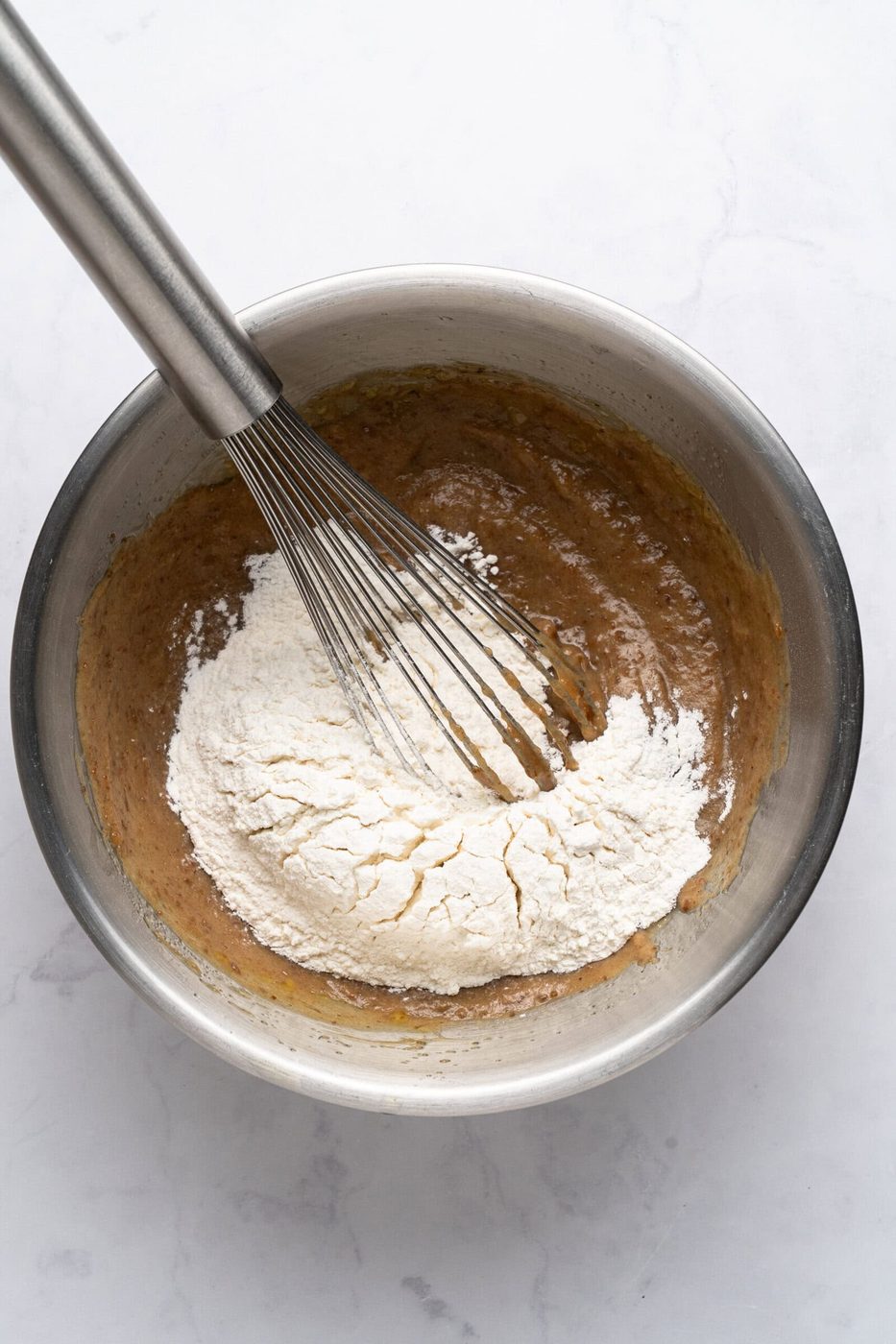 A metal bowl with a whisk mixing flour into batter. The mixture is partially combined, with some flour still visible on top, against a light marble background.