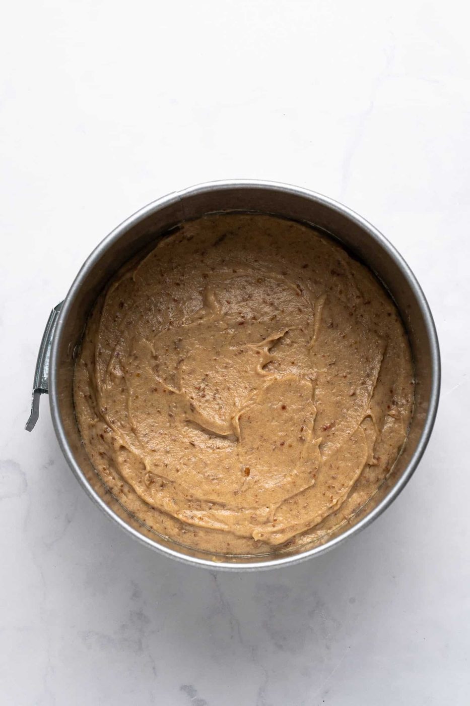 A round cake pan filled with smooth, light brown cake batter on a white marble surface.