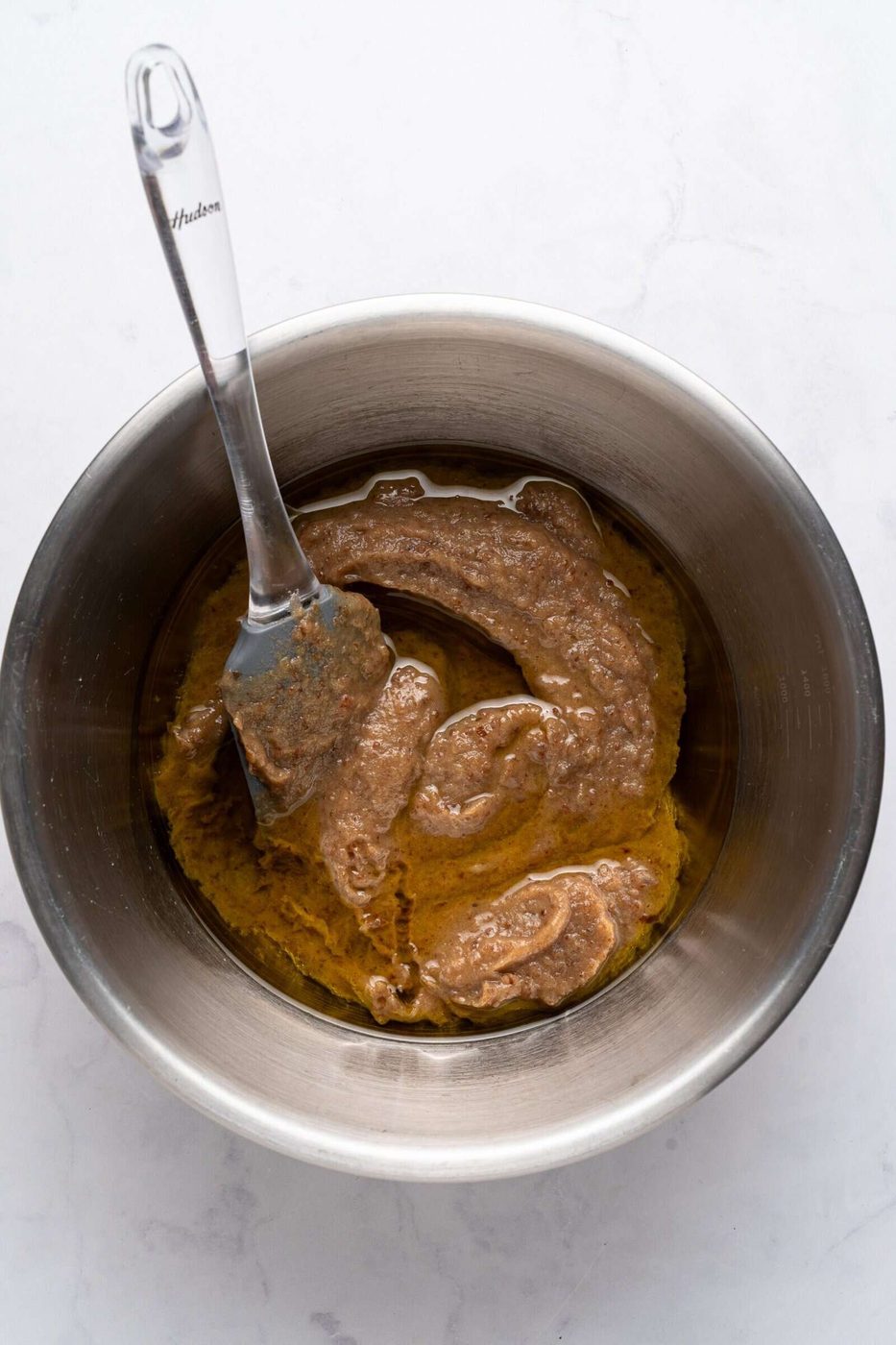 A stainless steel bowl on a light surface contains a mixture of nut butter and oil. A spatula with a transparent handle is partially submerged in the thick, brown mixture, which appears to have a smooth and slightly glossy texture.