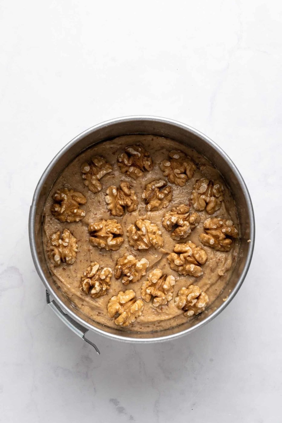 A round cake pan filled with light brown batter, topped with whole walnuts, sits on a light marble surface. The walnuts are evenly spaced on the surface of the batter, ready for baking.
