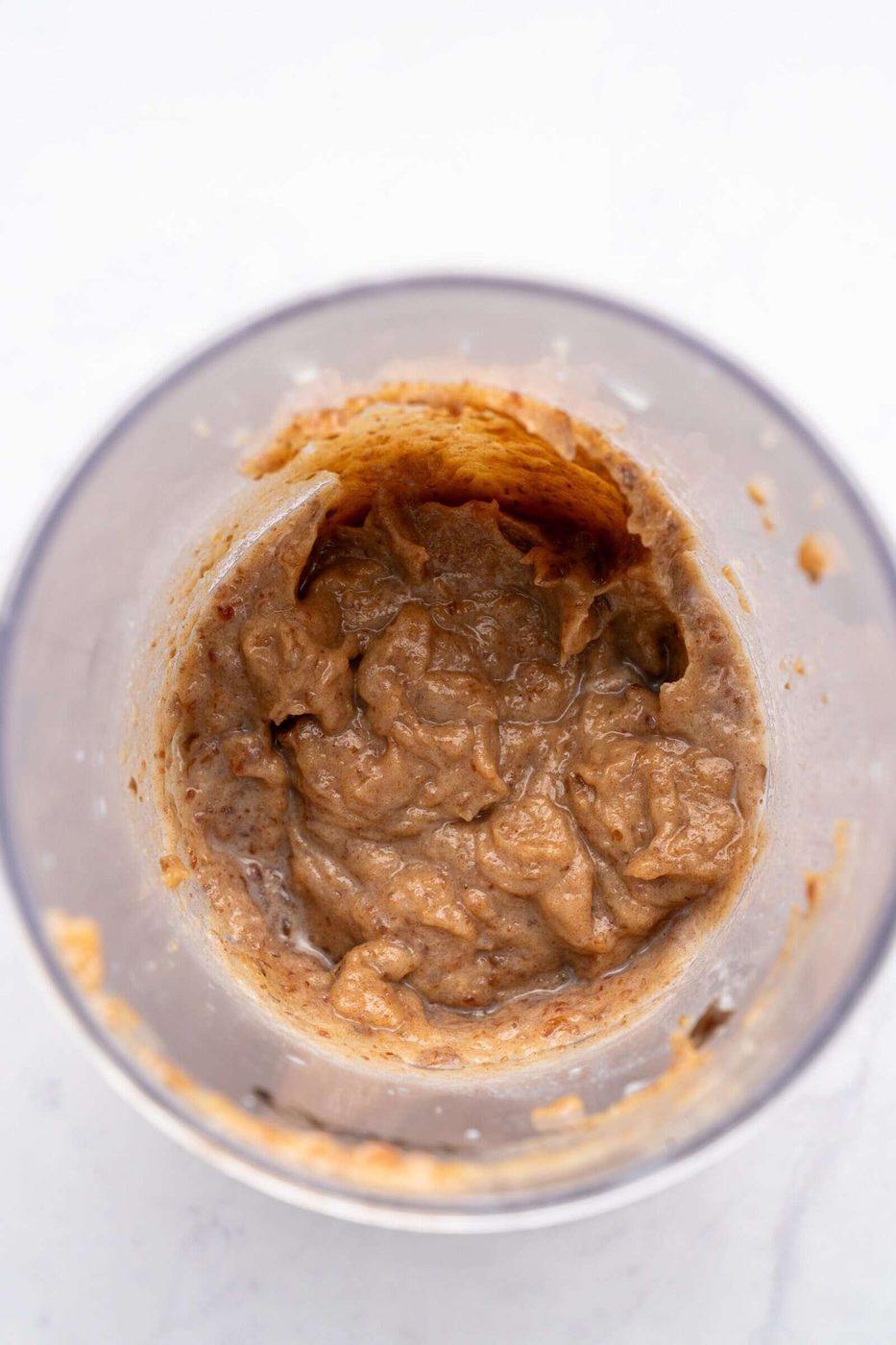 A close-up of a food processor bowl containing a blended brown mixture, likely a puree or paste, with a creamy texture. The sides of the bowl are partially coated with the mixture. The background is a light surface.