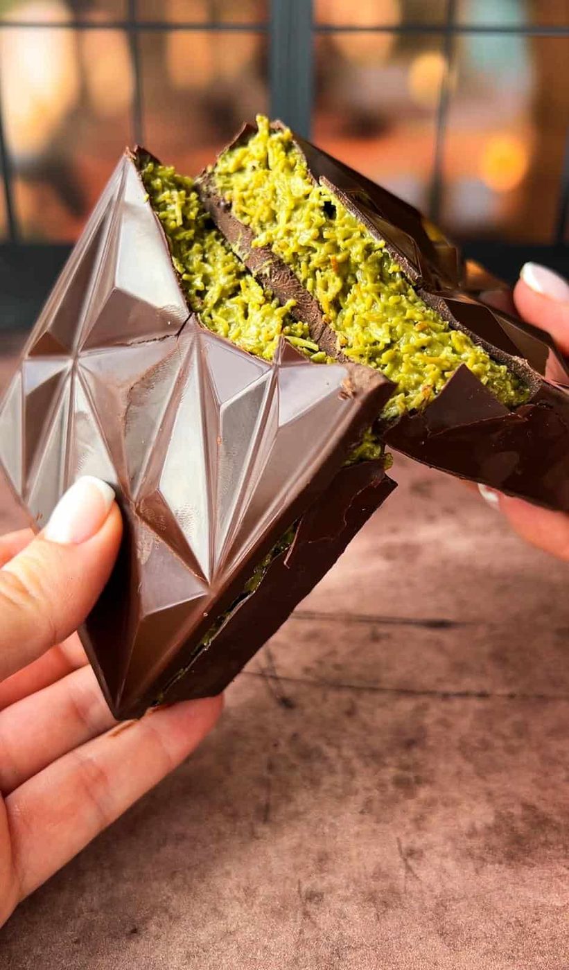 Two hands hold a chocolate dessert filled with a bright green, shredded filling. The chocolate has a geometric pattern, and the image is set against a blurred background with dark framing.