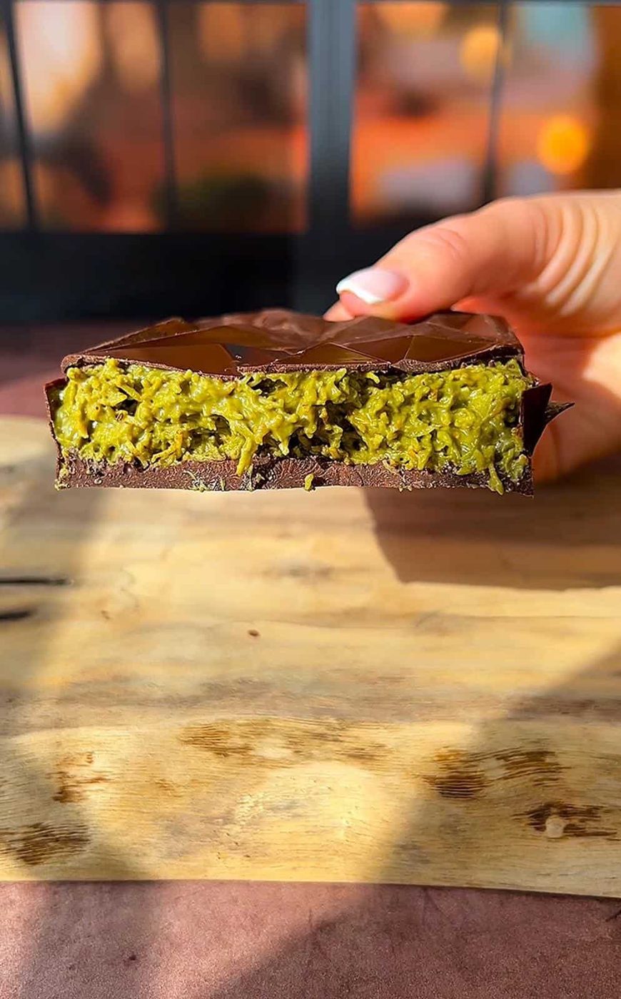 A close-up of a hand holding a cross-section of a chocolate bar with a green filling. The filling has a textured, fibrous appearance, and the bar is resting on a wooden surface with a blurred background.